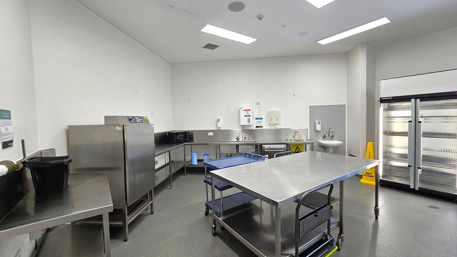 A commercial kitchen with stainless steel work surfaces and equipment. On the left, a black bin sits on a counter. In the centre, a large stainless steel table with wheels and a blue trolley. The back wall has a sink, microwave, and wall-mounted appliances. On the right, a large double-door refrigerator with a "Wet Floor" sign nearby. The floor is dark and speckled, and the ceiling has recessed lighting.