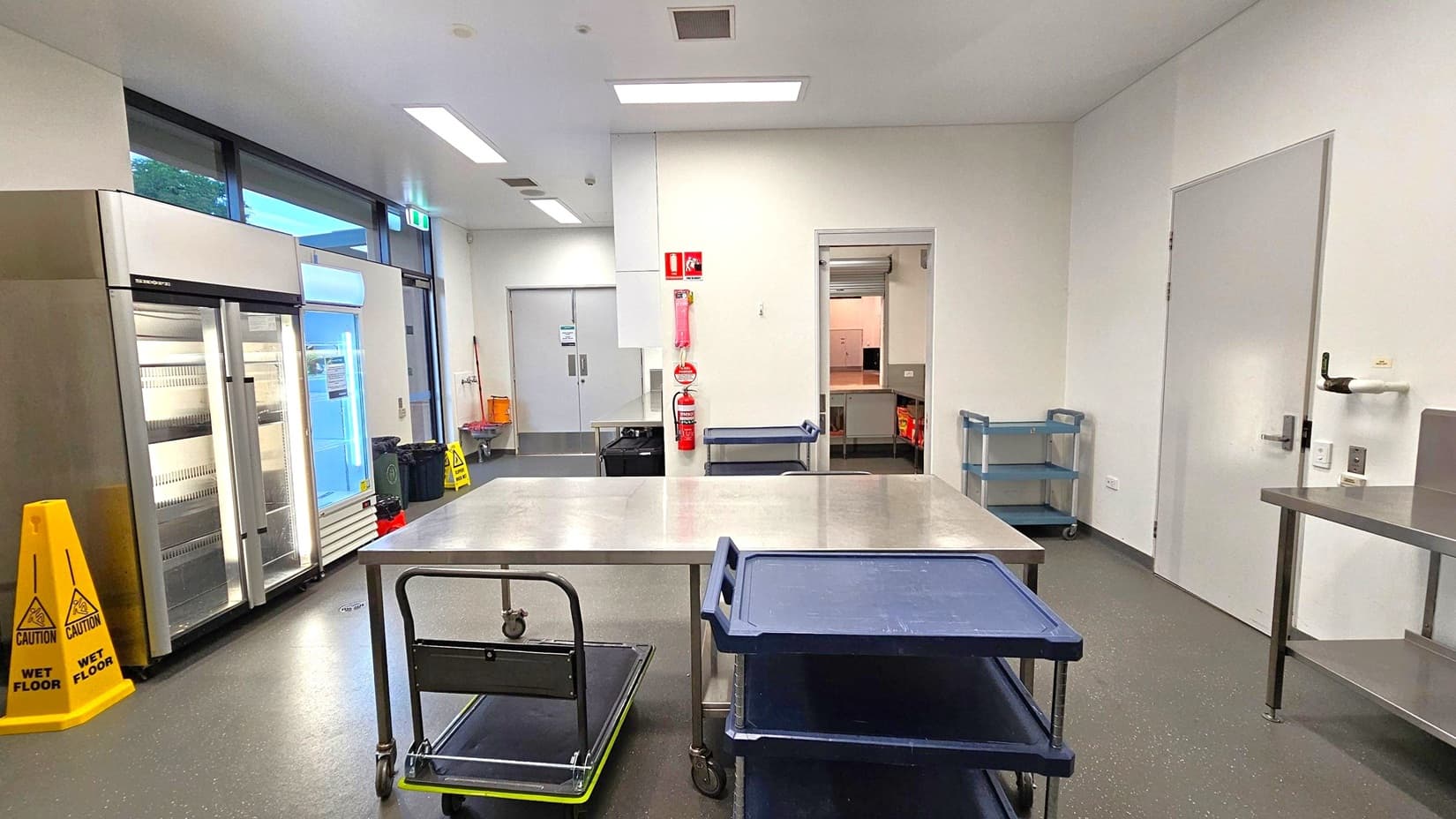 A commercial kitchen with a grey speckled floor and white ceiling. On the left, there are large glass-door refrigerators and a yellow caution sign. In the centre, a stainless steel table is surrounded by trolleys. The back wall has a fire extinguisher and two doors, one leading to another room. On the right, a wall-mounted paper towel dispenser and a metal shelf are visible. The room is well-lit with fluorescent lights.