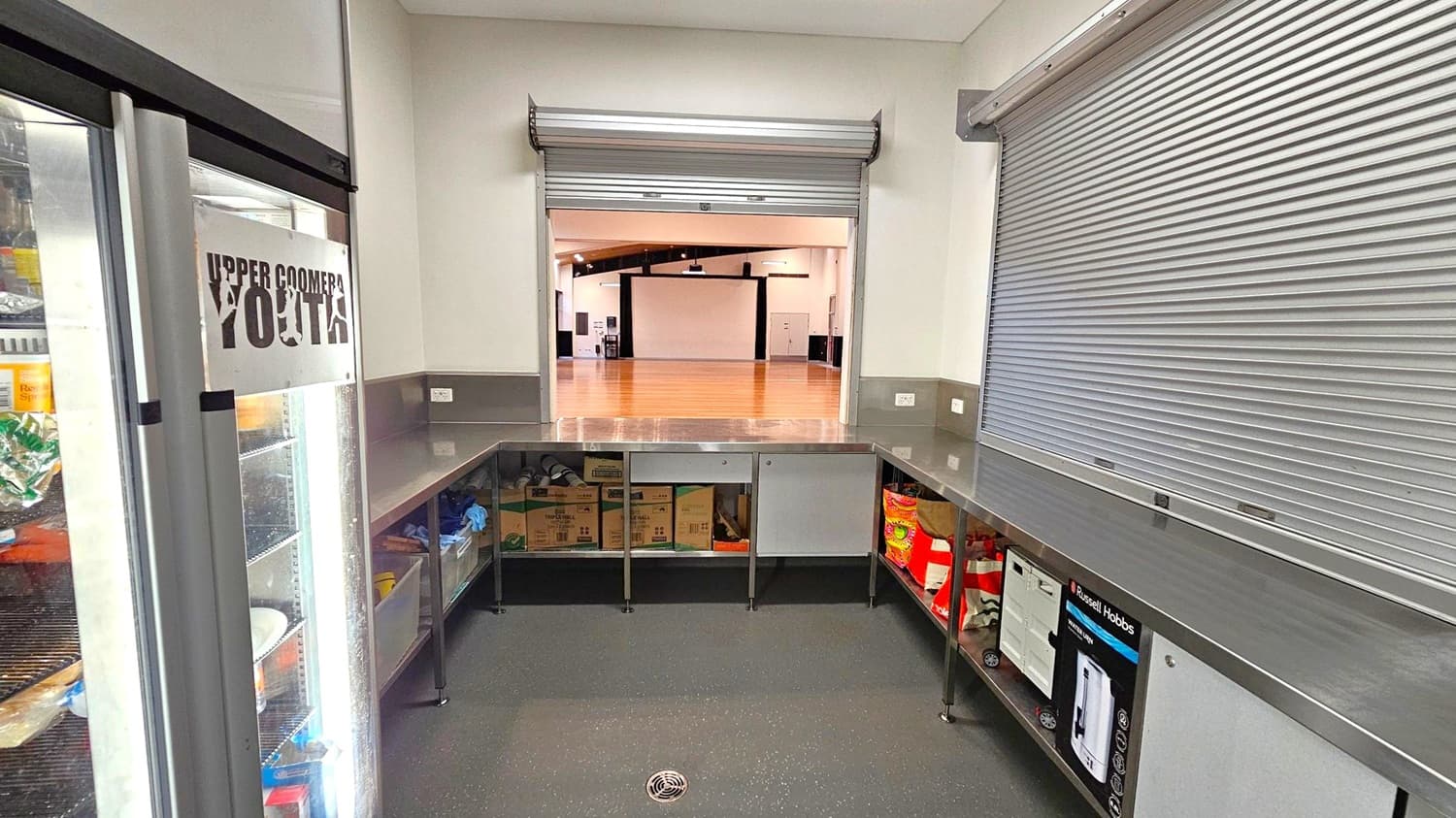 A small kitchen area with stainless steel countertops and open shelving underneath, containing various boxes and items. On the left, a glass-fronted refrigerator displays food items, with a sign reading "Upper Coomera Youth." In the centre, a partially open roller shutter reveals a large, empty hall with wooden flooring. On the right, another closed roller shutter is visible. The floor is dark with a drain in the centre. The ceiling is plain and light-coloured.