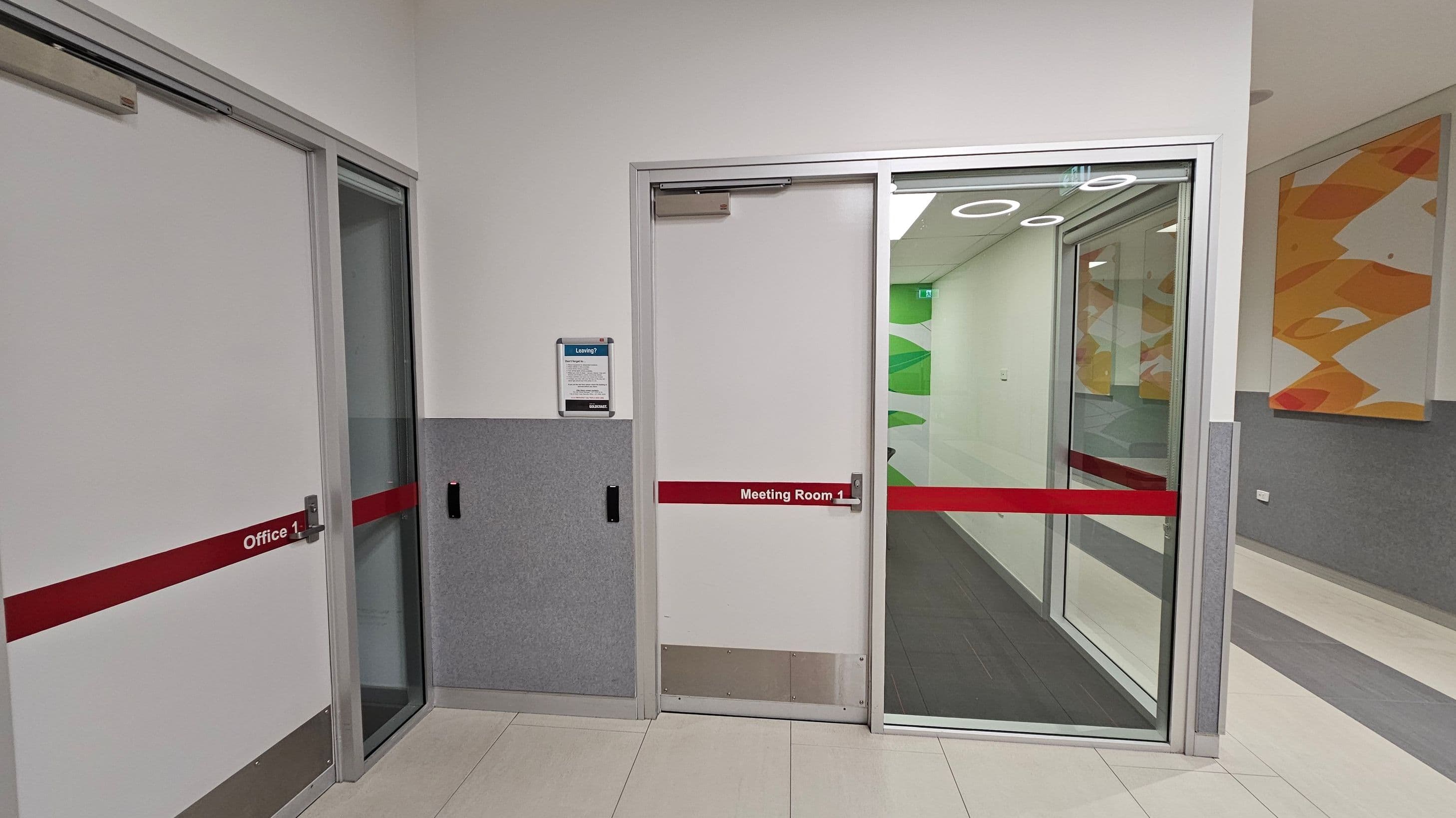 A hallway with two doors on the left side. The first door is labelled "Office 1" and the second "Meeting Room 1," both with red horizontal stripes. The meeting room door has a glass panel showing a corridor with green and white wall designs. A notice board is mounted on the wall between the doors. The floor is tiled in light colours, and the walls are grey and white. On the right, there is a colourful abstract painting.
