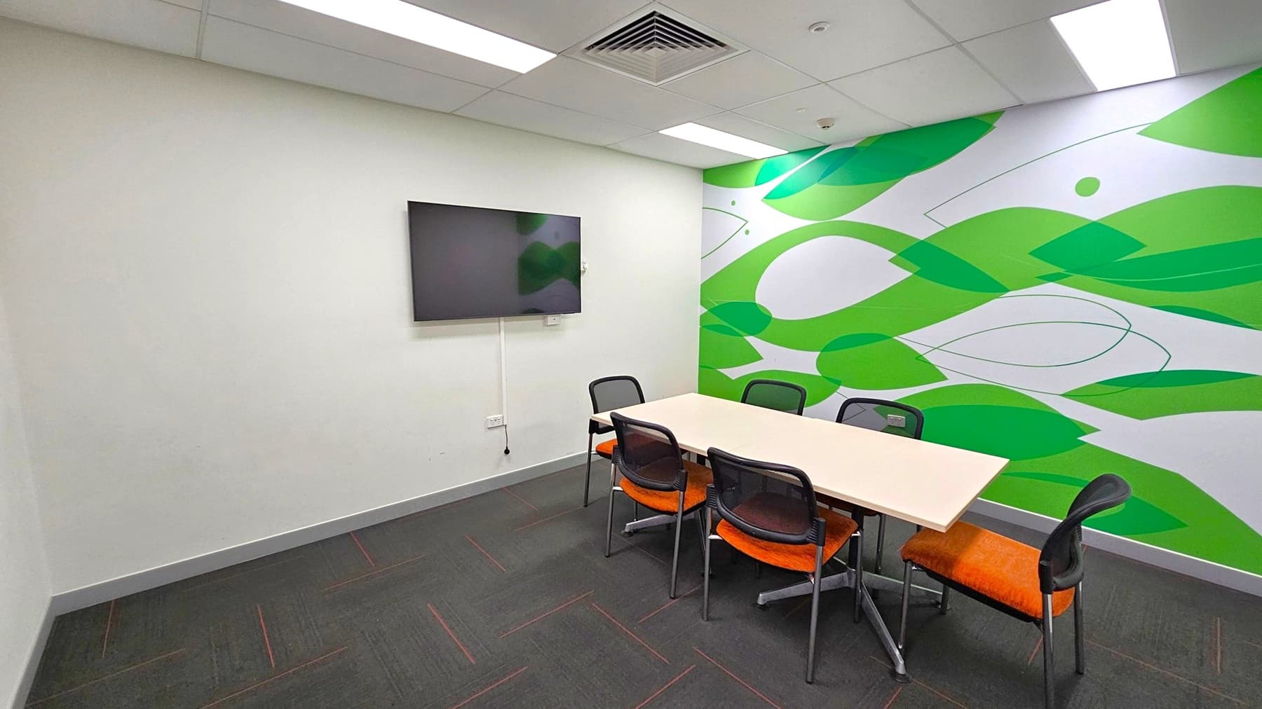 A small meeting room with a rectangular table surrounded by five chairs with orange seats. The right wall features a vibrant green and white abstract design. On the left wall, a flat-screen TV is mounted. The floor is dark with subtle red lines, and the ceiling has recessed lighting and a central air vent.