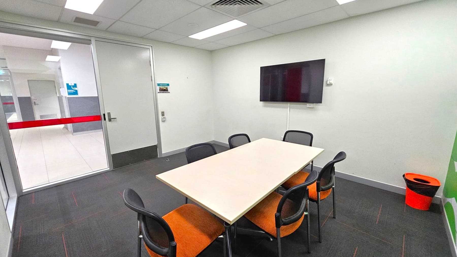 A small meeting room with a rectangular table surrounded by six chairs with orange seats. On the right wall, a flat-screen TV is mounted. A bright orange bin is placed in the corner. The left side features a glass door with a red stripe, leading to a corridor. The floor is carpeted in dark grey, and the ceiling has recessed lighting and an air vent. Walls are plain white with minimal decoration