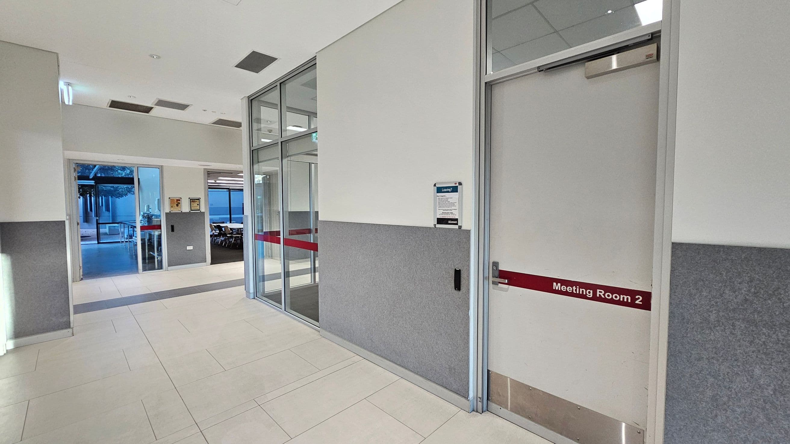 A corridor with light-coloured tiled flooring and a white ceiling with recessed lights. On the right, a door labelled "Meeting Room 2" with a red stripe. Adjacent to it, a grey and white wall with a notice board. On the left, a glass door leads to an outdoor area with visible trees and a table. Further inside, another room with chairs and tables is visible through a glass partition.