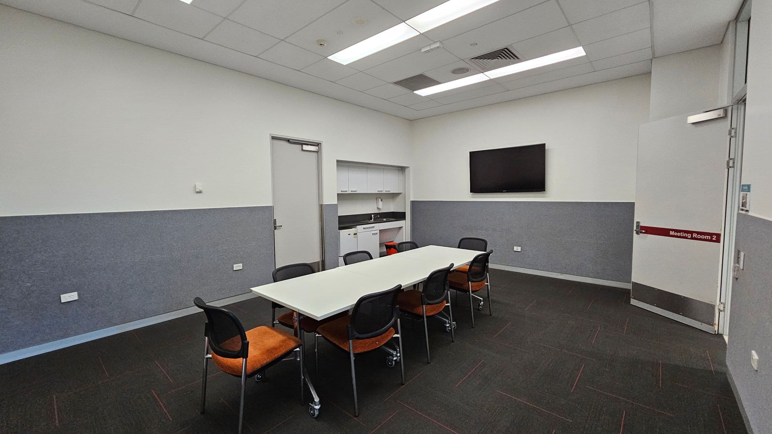 Meeting room with a rectangular white table in the centre surrounded by six chairs with orange seats. On the right wall, a flat-screen TV is mounted. Below the TV, a grey panel runs along the wall. A door labelled "Meeting Room 2" is on the far right. On the left, a door leads to a small kitchenette area with cabinets and a sink. The ceiling has recessed lighting and a grid pattern. The floor is carpeted in dark grey.