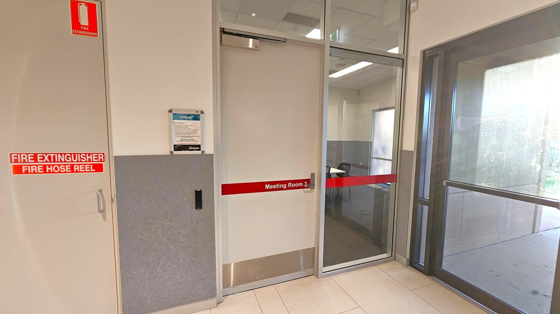 A meeting room with a rectangular white table in the centre surrounded by six chairs with orange seats. On the right wall, a flat-screen TV is mounted. Below the TV, a door labelled "Meeting Room 2" is visible. On the left, there is a door and a small kitchenette with cabinets and a sink. The walls are white with a grey lower section. The ceiling has recessed lighting and ventilation panels. The floor is carpeted in dark grey.