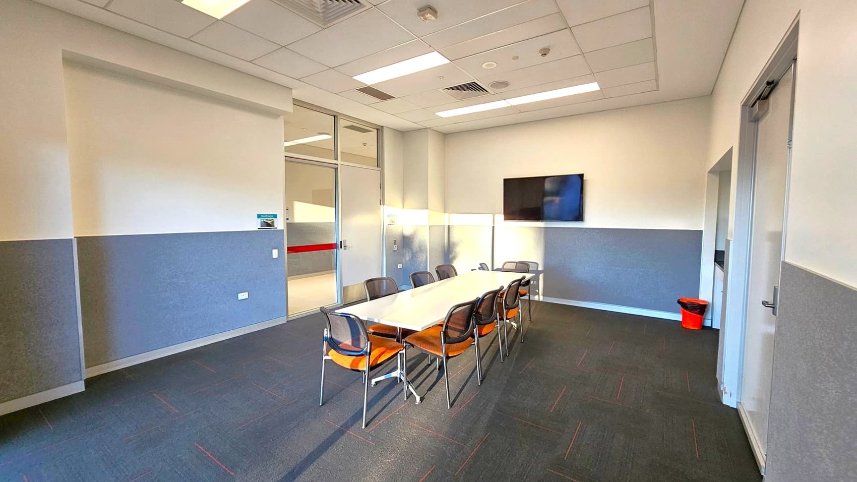 A corridor with a door labelled "Meeting Room 3" in the centre, featuring a red stripe. To the left, a cabinet with signs for a fire extinguisher and fire hose reel. A notice board is mounted on the wall above. On the right, a large glass window provides a view outside. The floor is tiled in white, and the ceiling has recessed lighting.