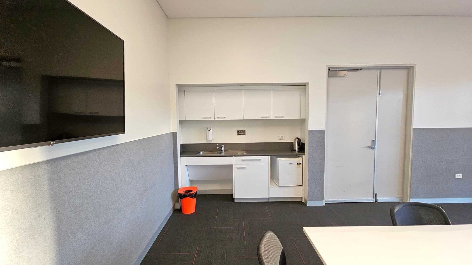 A conference room with a long white table in the centre surrounded by eight chairs with orange seats. On the right wall, a flat-screen TV is mounted above a grey panel. A bright red bin is placed in the corner. The left side features a glass door with a red stripe, leading to another room. The walls are painted white with grey panelling, and the ceiling has recessed lighting and ventilation panels. The floor is carpeted in a dark grey tone.