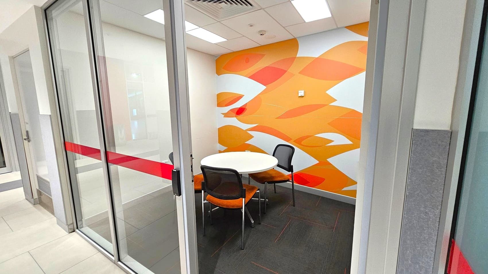 Small meeting room with glass sliding doors featuring a red stripe. Inside, a round white table is surrounded by three chairs with orange seats and black backs. The back wall is decorated with an abstract orange and white pattern. The floor is dark grey, and the ceiling has recessed lighting and air vents.