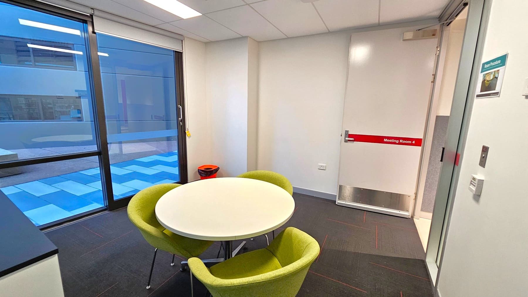 A small meeting room with a round white table surrounded by three green chairs on a dark tiled floor. To the right, a door labelled "Meeting Room 4" is slightly open. On the left, large windows with blue-tinted glass overlook an outdoor area with a patterned floor. A small orange bin is placed in the corner near the window. The ceiling has recessed lighting.