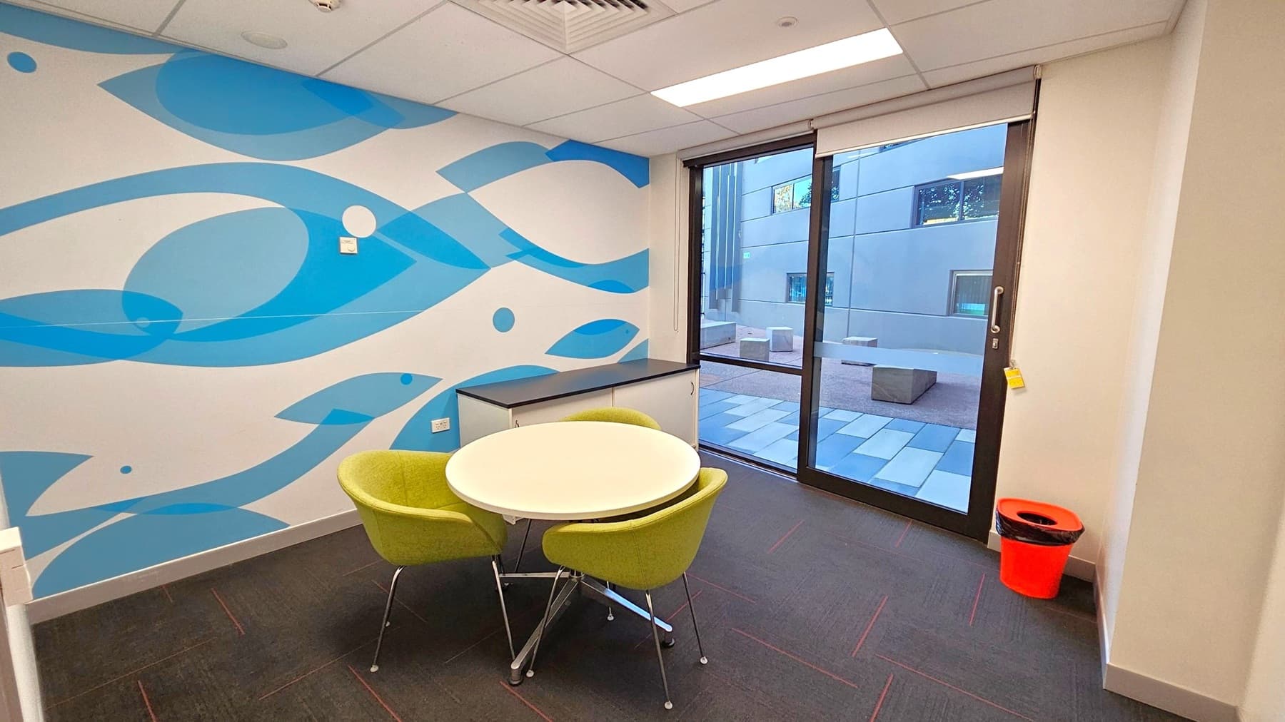 A small room with a white ceiling and dark floor. On the left, a wall features a blue abstract pattern. In the centre, a round white table is surrounded by three green chairs. To the right, a large glass door leads to an outdoor area with a tiled floor and concrete seating. A bright orange bin is placed near the door. The room has a modern and minimalistic design.