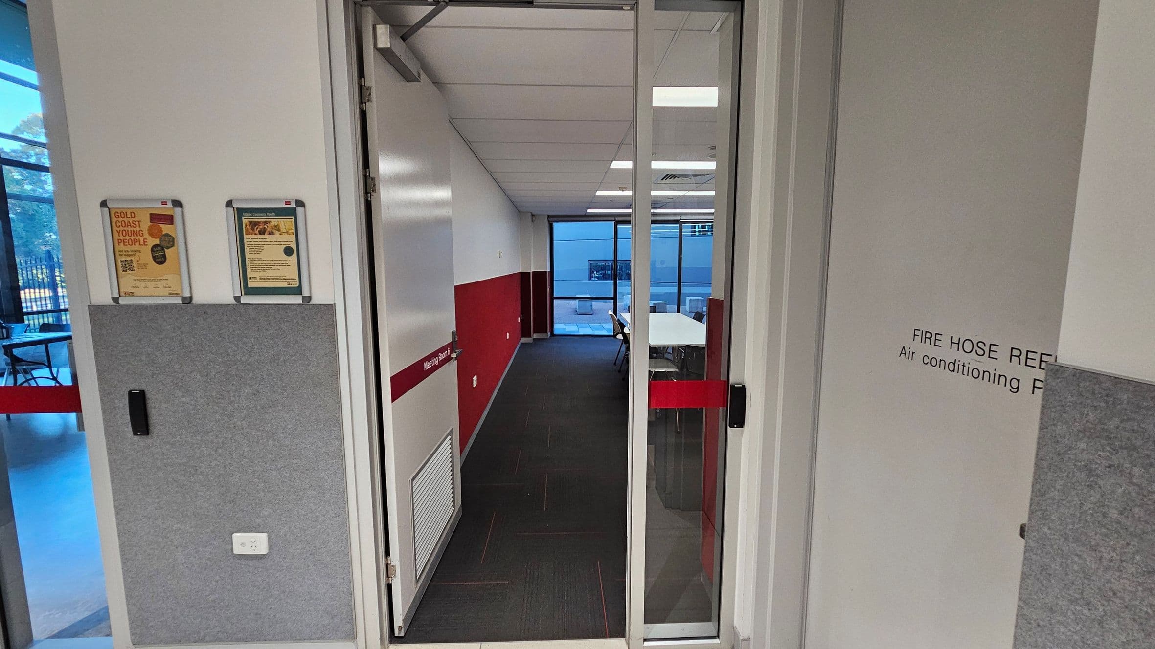 A corridor leading to a meeting room with a glass door. The walls are white with a red stripe running horizontally. On the left, there are two framed notices on the wall above a grey panel. The floor is carpeted in dark grey. In the background, the meeting room has a table and chairs, with large windows showing an outdoor area. The ceiling is fitted with white panels and fluorescent lights. On the right, there is a sign indicating a fire hose reel and air conditioning panel.