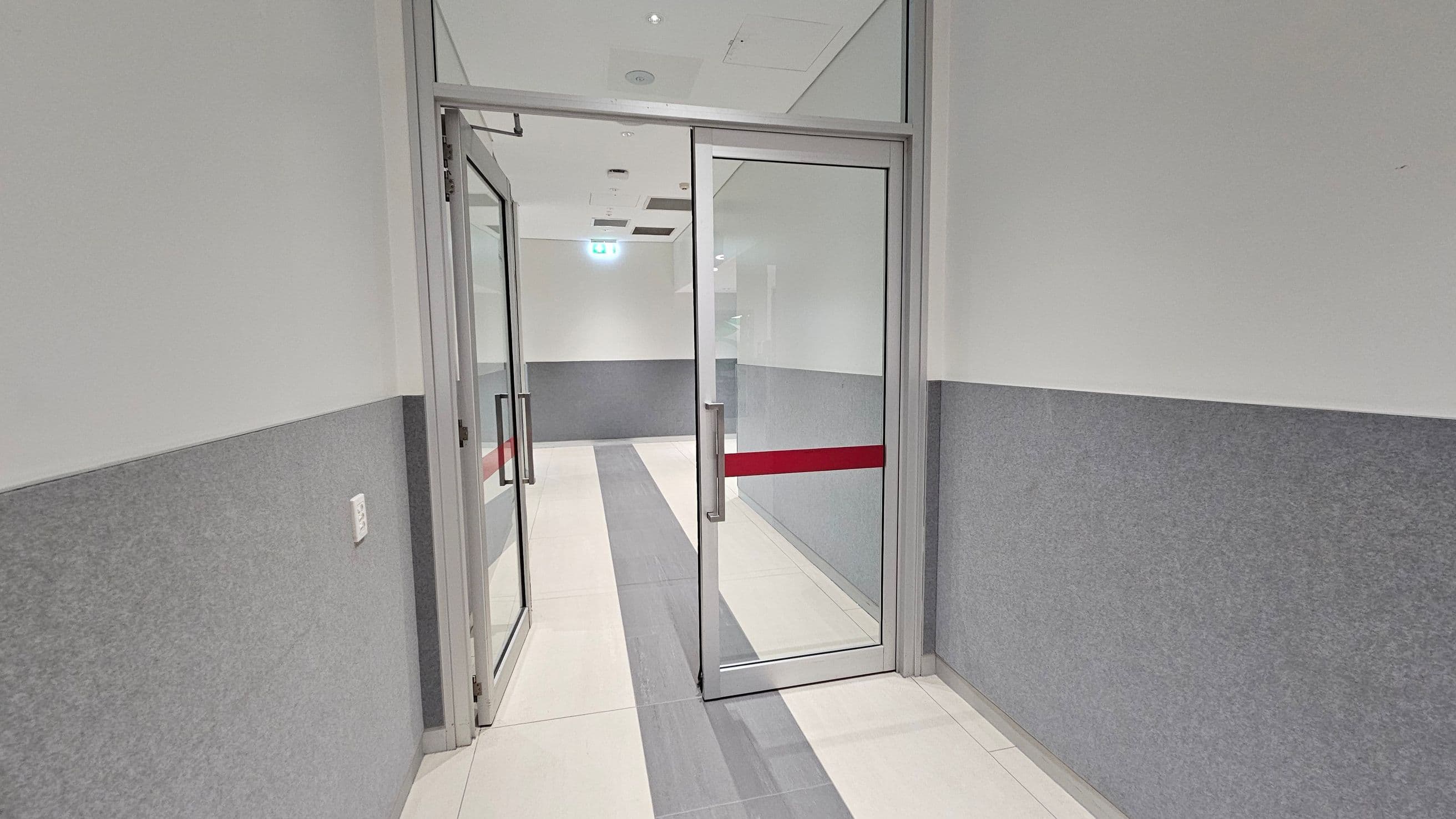 A corridor leading to a meeting room with a glass door. The walls are white with a red stripe running horizontally. On the left, there are two framed notices on the wall above a grey panel. The floor is carpeted in dark grey. In the background, the meeting room has a table and chairs, with large windows showing an outdoor area. The ceiling is fitted with white panels and fluorescent lights. On the right, there is a sign indicating a fire hose reel and air conditioning panel.