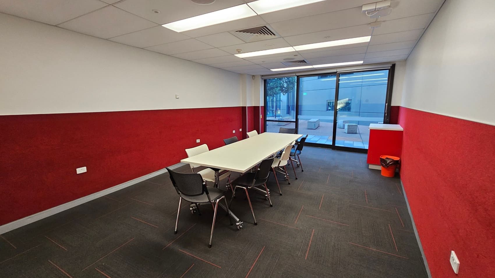 A meeting room with a long white table in the centre surrounded by black and white chairs on a dark carpeted floor. The walls are painted red and white, with a large glass door on the right leading to an outdoor area. A small red cabinet with an orange bin is in the corner near the door. The ceiling is fitted with fluorescent lights and air vents.