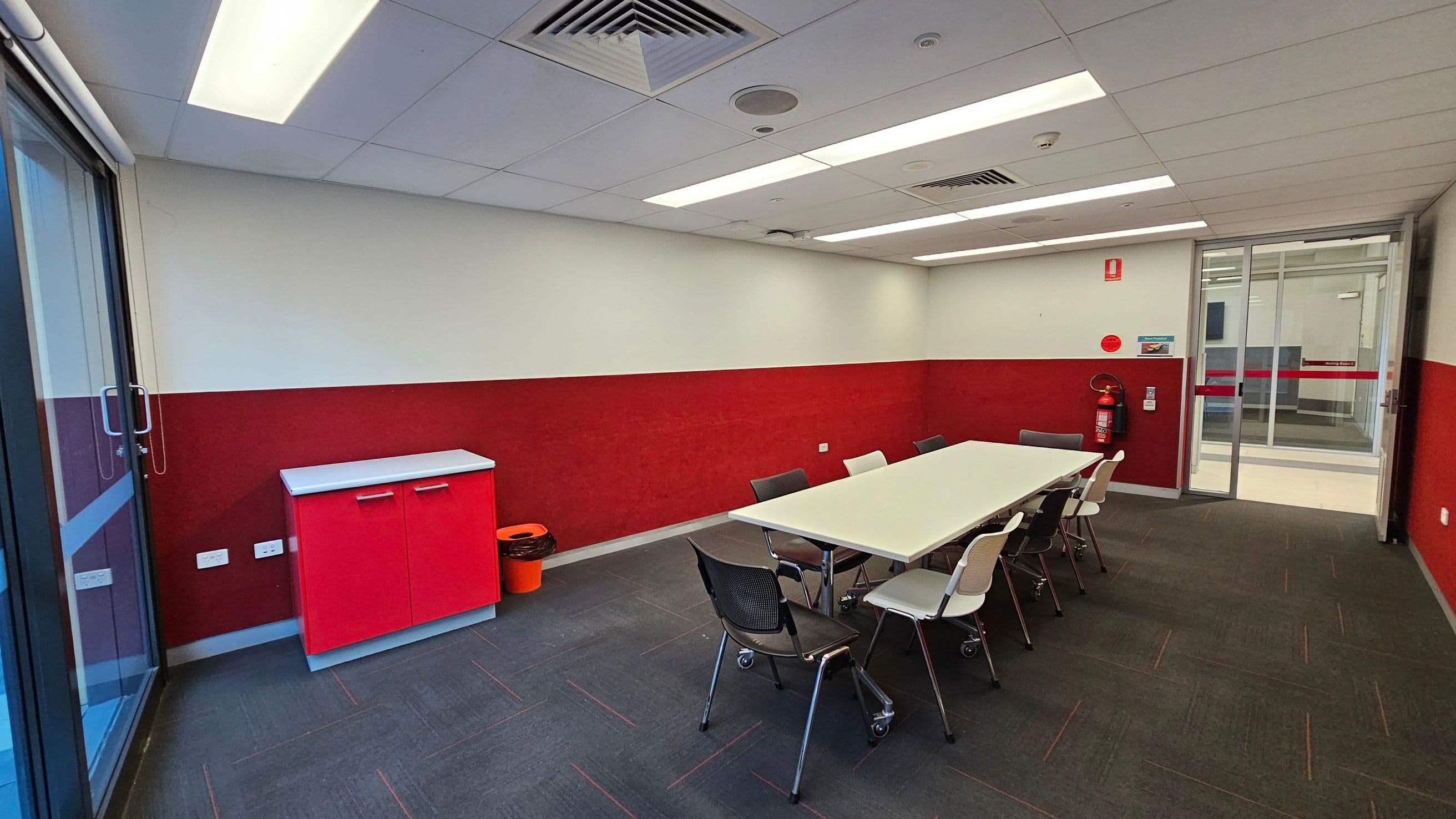 A room with a red and white colour scheme. On the left, there's a glass door and a red cabinet with a white top. An orange bin is next to the cabinet. In the centre, a white rectangular table is surrounded by black and white chairs. On the right, a fire extinguisher is mounted on the wall near a glass door. The floor is carpeted in dark grey, and the ceiling has recessed lighting and air vents.