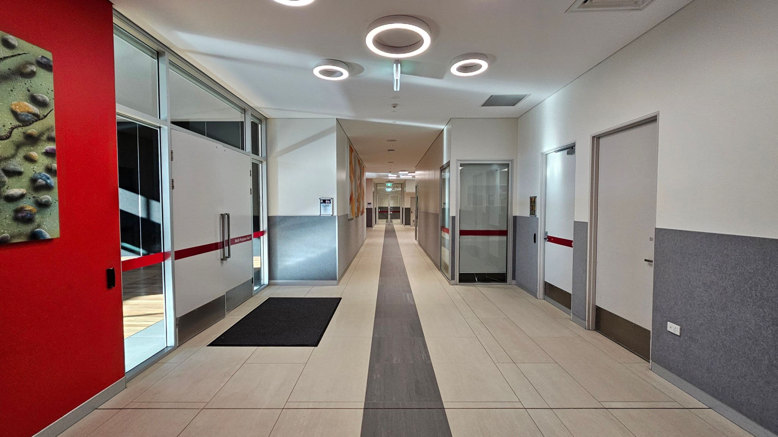 A modern hallway with a light-coloured tiled floor and grey accents. On the left, there's a red wall with a textured artwork and glass doors with red stripes. On the right, there are several doors with grey and white walls. The ceiling features circular light fixtures. The hallway extends into the distance with more doors and lights visible.