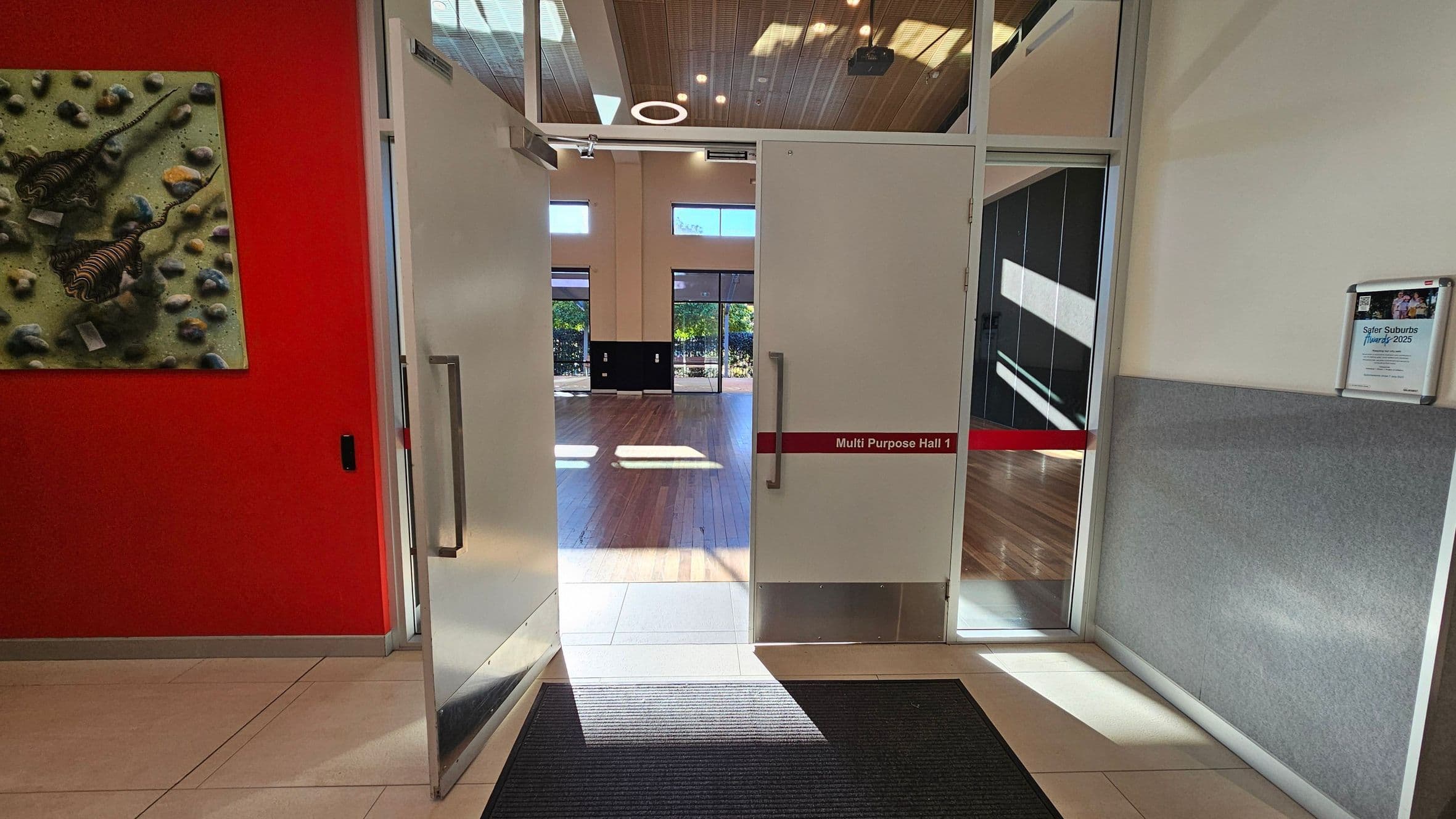 Double doors open into a spacious hall with wooden flooring. The left wall is red with a textured artwork featuring abstract shapes. The right wall is light-coloured with a grey panel and a notice board displaying a poster. Above, the ceiling is wooden with circular lights. The floor in the foreground is tiled with a dark mat near the entrance. The hall has large windows at the far end, allowing natural light to illuminate the space.