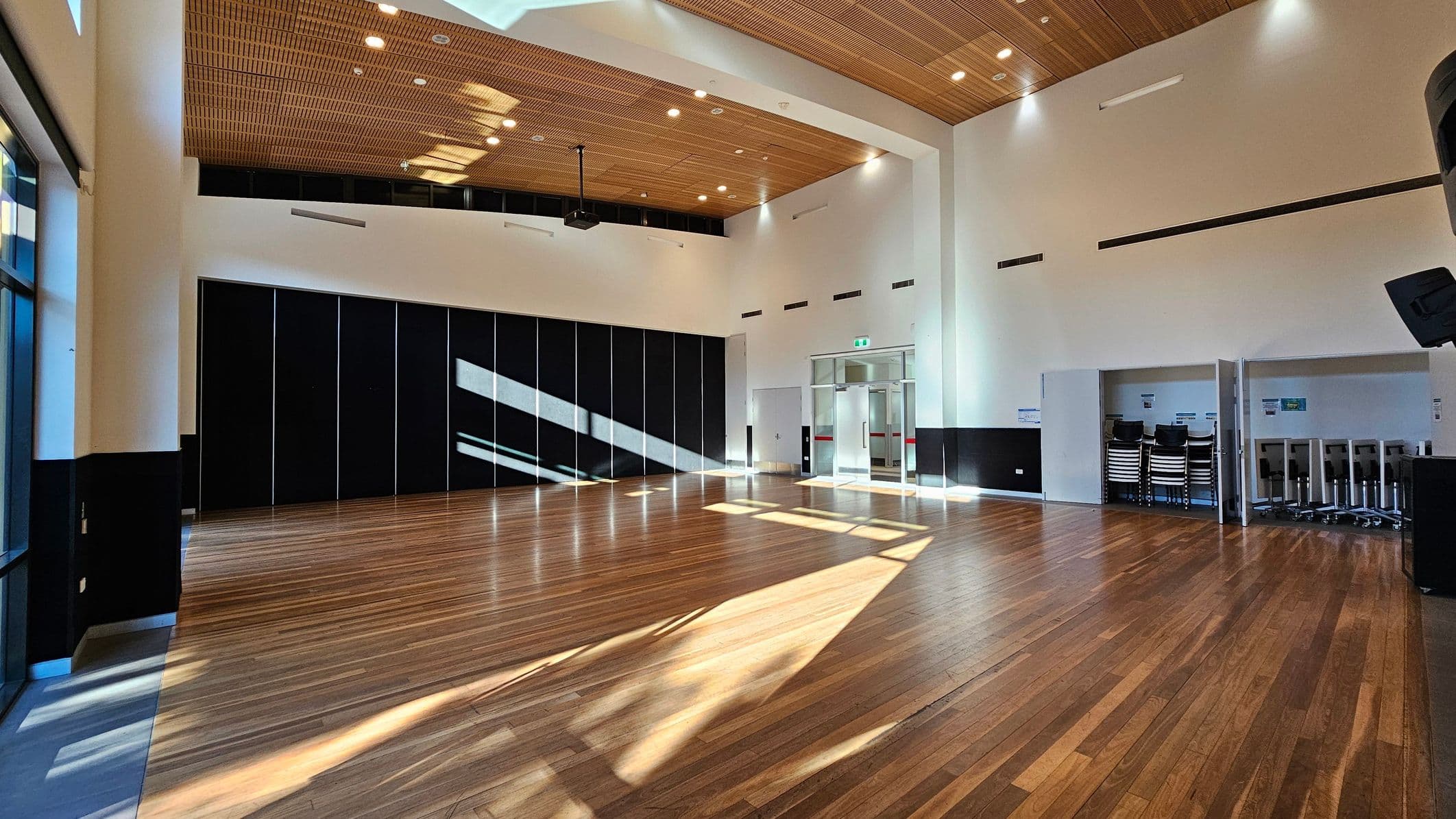 A spacious room with a wooden floor and a high ceiling featuring wooden panels. On the left, large windows allow natural light to create patterns on the floor. The back wall has black panels, and a projector is mounted on the ceiling. On the right, there are stacked chairs and tables stored in an alcove. The room has white walls with black accents and recessed lighting. A glass door is visible in the centre of the back wall.