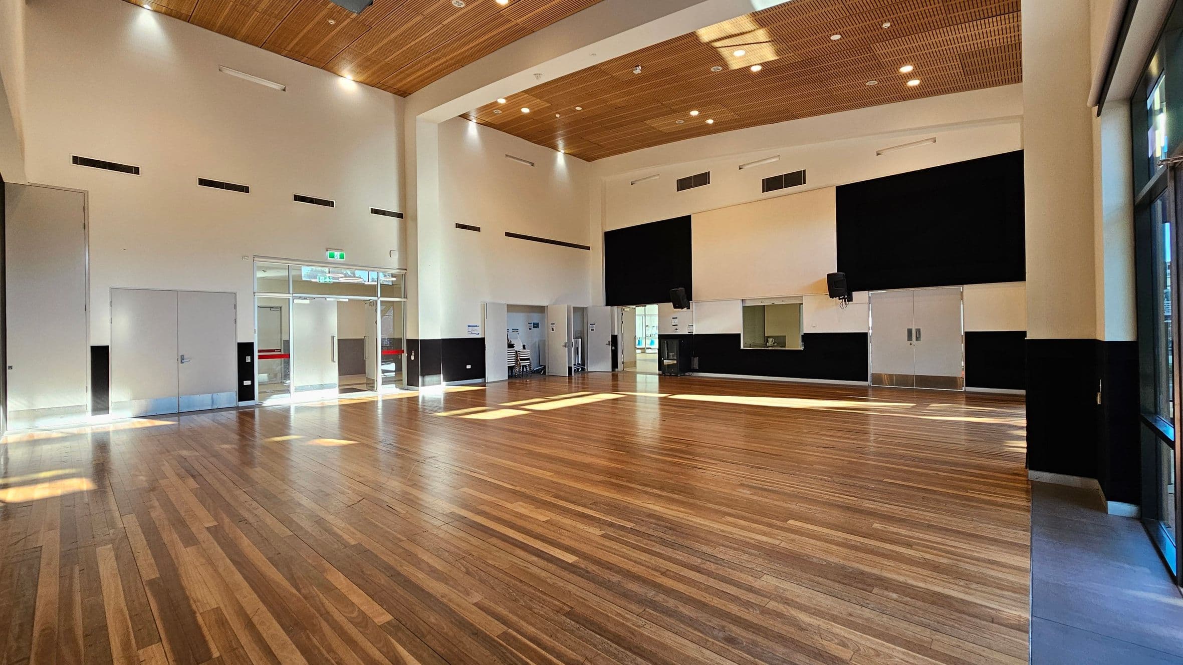 A spacious hall with a polished wooden floor and high ceiling. The ceiling features wooden panelling with recessed lighting. On the left, there are large double doors and a glass entrance. The right side has a wall with large black panels and a service window. Sunlight streams through the windows, creating patterns on the floor. The walls are mostly white with black accents at the bottom.