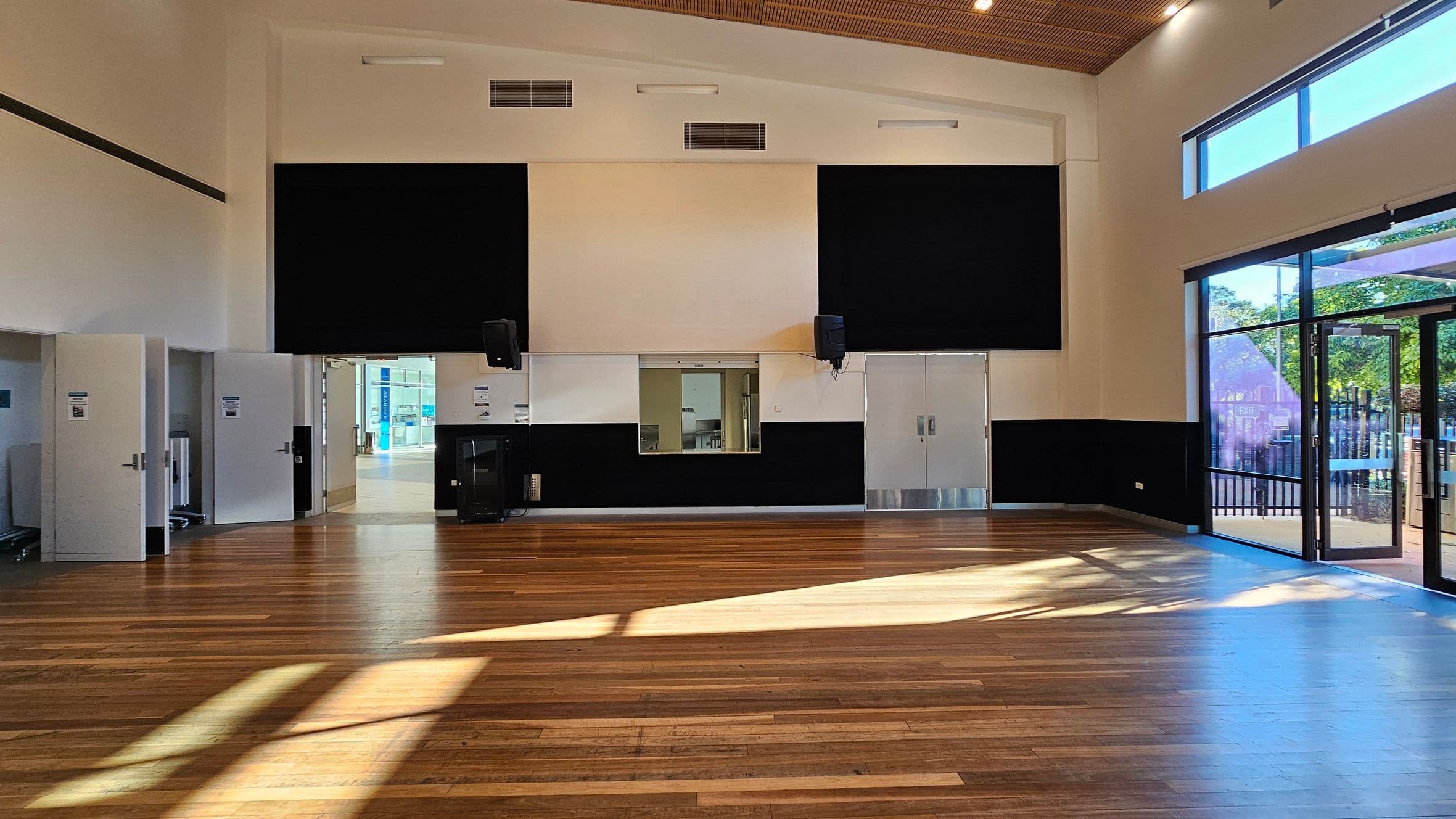 A spacious room with wooden flooring and a high ceiling. On the left, there are open doors leading to another area. In the centre, a wall features two large black panels with speakers mounted on them, and a small window in between. On the right, large glass doors and windows allow natural light to flood in, casting shadows on the floor. Outside, greenery is visible through the glass. The ceiling has recessed lighting and wooden panelling.