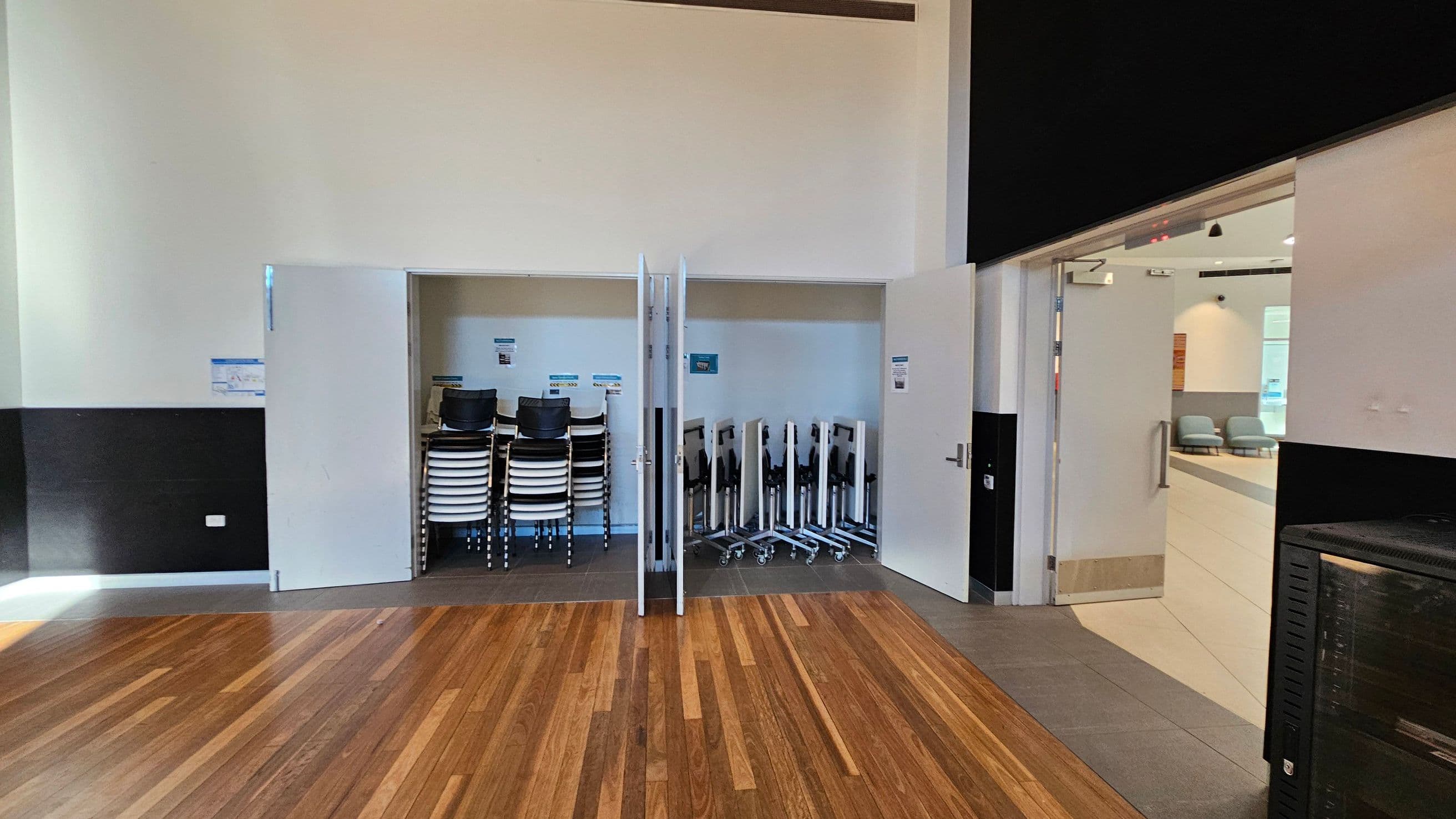 A room with a wooden floor and a high ceiling. In the centre, there's a storage area with stacks of chairs and tables on wheels. To the right, an open door leads to another room with light-coloured walls and seating. The walls are mostly white with a black section at the bottom. A black cabinet is visible on the right side.
