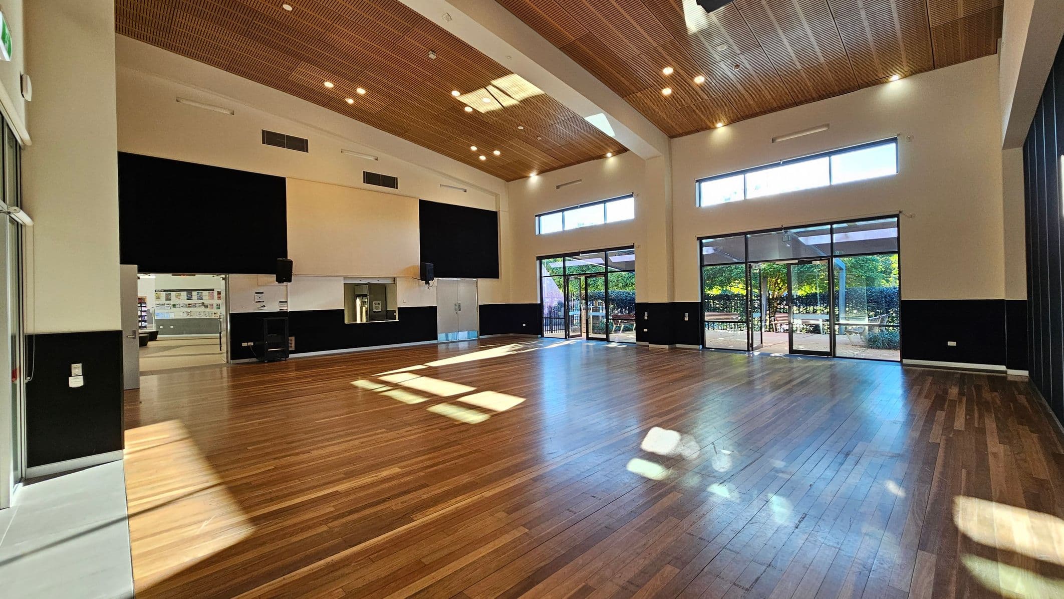 A spacious room with polished wooden flooring and a high ceiling featuring wooden panels. Large windows and glass doors on the right allow natural light to flood in, revealing an outdoor area with greenery. On the left, there is a doorway leading to another room. The walls are painted in a light colour with black accents. Ceiling lights are evenly distributed, and the room appears to be a multi-purpose space, possibly for events or gatherings.