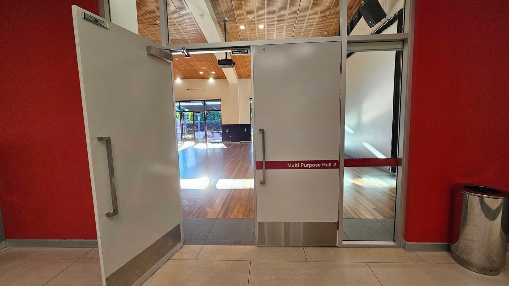 Double doors open into a spacious hall with a wooden floor and a high ceiling featuring wooden panels and recessed lighting. The doors are labelled "Multi Purpose Hall 2" and are set in a red wall. To the right, a shiny metal bin is visible. The hall has large windows on the far side, allowing natural light to stream in, and a projector is mounted on the ceiling. The floor outside the hall is tiled.