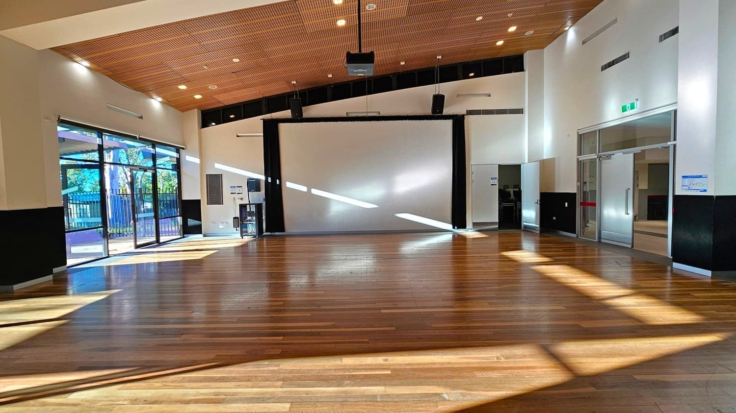 A spacious room with a wooden floor and a high ceiling featuring recessed lights. On the left, large glass doors and windows allow natural light to stream in, casting shadows on the floor. In the centre, a large white screen is mounted on the wall. On the right, there are glass doors with metal frames and an emergency exit sign above. The walls are painted white with a black lower section.