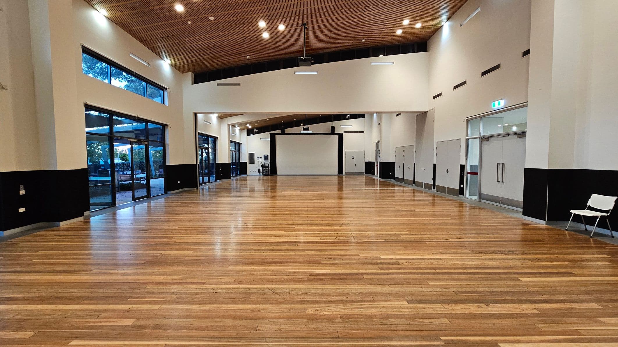 A spacious hall with a polished wooden floor and a high, wooden-panelled ceiling. On the left, large glass windows and doors allow natural light in, with views of trees outside. On the right, there are several doors and a single white chair. At the far end, a large white screen is mounted on the wall. The ceiling features multiple recessed lights, and the walls are painted in a light colour with a dark band running along the lower section.