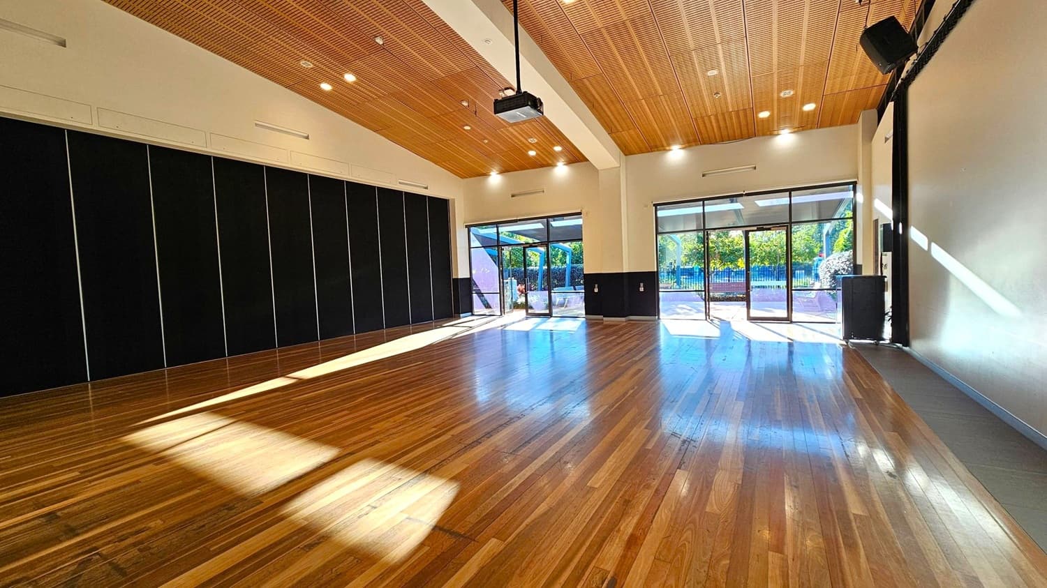 A spacious room with a polished wooden floor and a wooden panelled ceiling. On the left, there are large black panels or partitions. The right side features a plain wall with a speaker mounted near the ceiling. In the centre, large glass doors and windows allow natural light to flood in, revealing a view of greenery outside. A projector is mounted on the ceiling. The room is well-lit with ceiling lights.