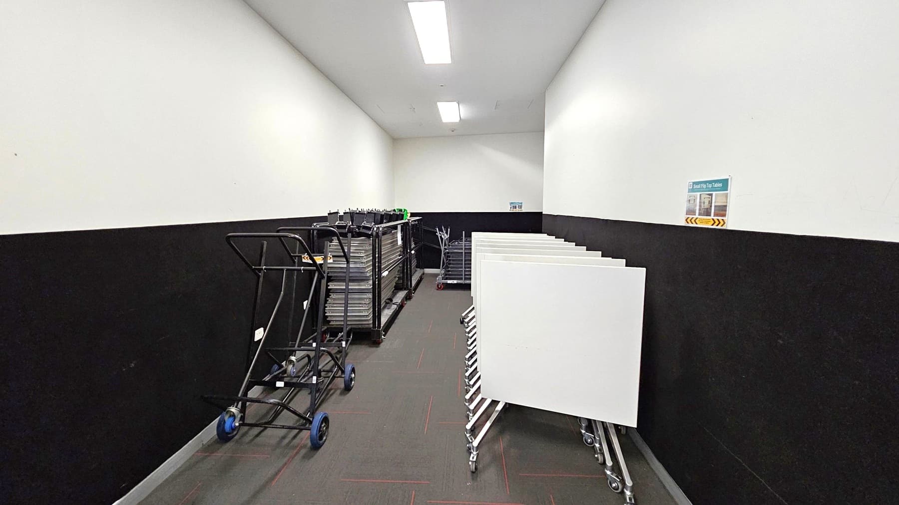 A narrow storage room with a grey floor and white ceiling. On the left, there are black trolleys with blue wheels and stacks of trays. On the right, several white boards are stored on wheeled stands. The walls are painted white with a black lower half. A small sign is visible on the right wall. The room is lit by rectangular ceiling lights.