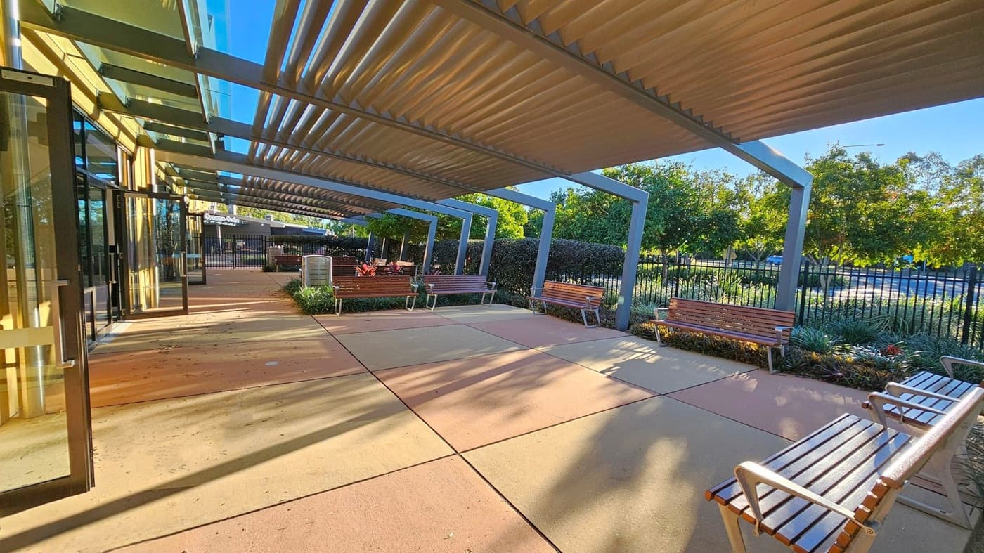 A covered outdoor area with a series of metal beams and slatted roofing providing shade. On the left, there is a glass entrance to a building. The ground is paved with large, square tiles. Wooden benches are placed along the right side, facing a fence with greenery and trees beyond it. In the background, more benches and tables are visible, suggesting a seating area. The scene is well-lit, indicating a sunny day.