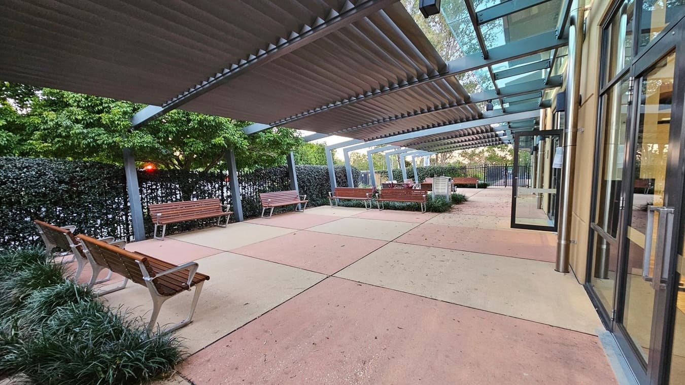 A covered outdoor seating area with several wooden benches on a paved surface. On the left, there are green plants and a hedge providing a natural backdrop. The right side features a building with large glass windows and doors. Above, a metal and glass canopy provides shade. The area is open and spacious, with a mix of concrete and tiled flooring.