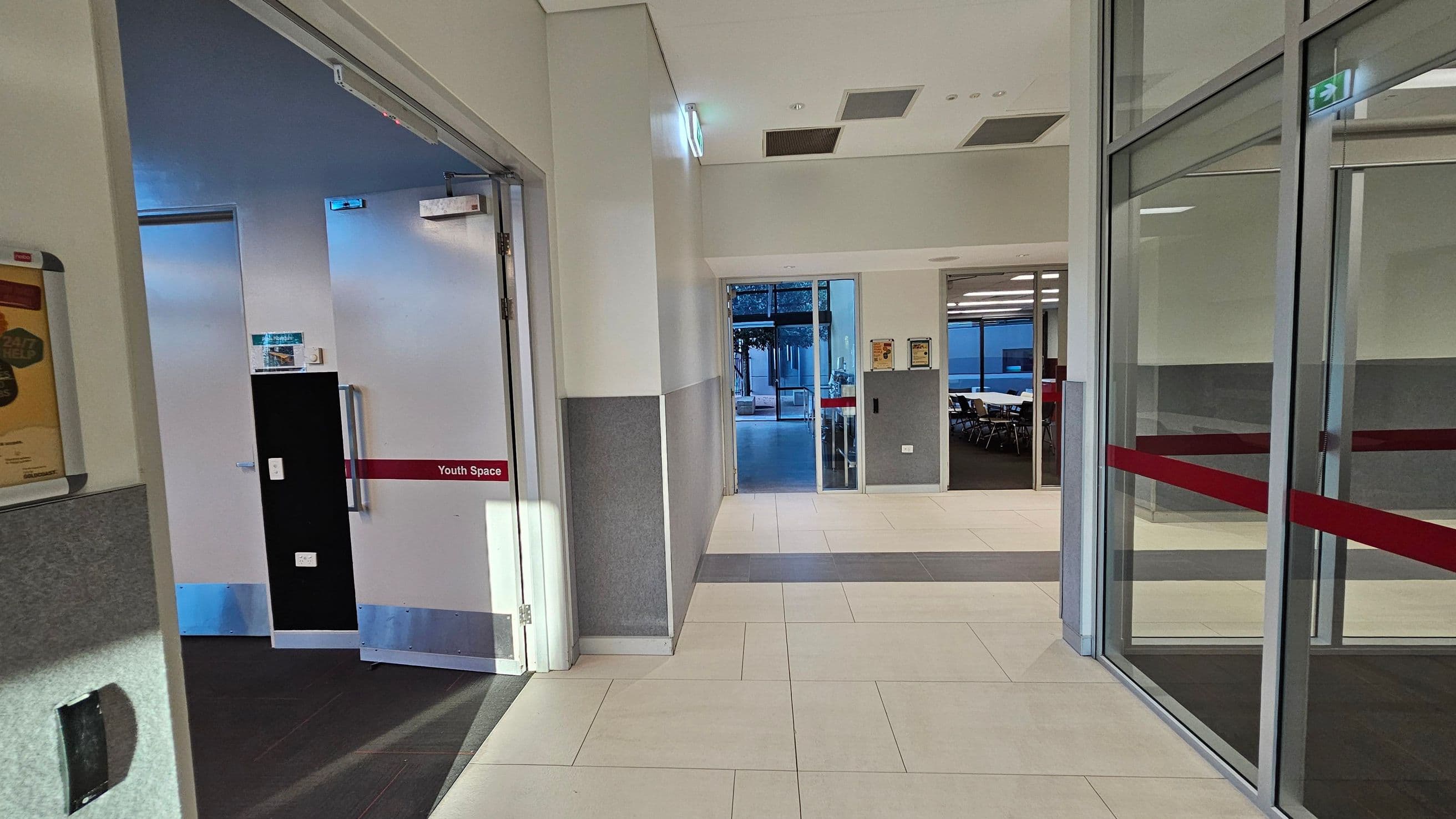 A hallway with light-coloured tiled flooring and grey walls. On the left, a door marked "Youth Space" is open, revealing a dark interior. In the centre, a glass door leads to an outdoor area with visible trees. On the right, a glass wall with red stripes separates another room with tables and chairs. The ceiling has recessed lighting and ventilation panels.