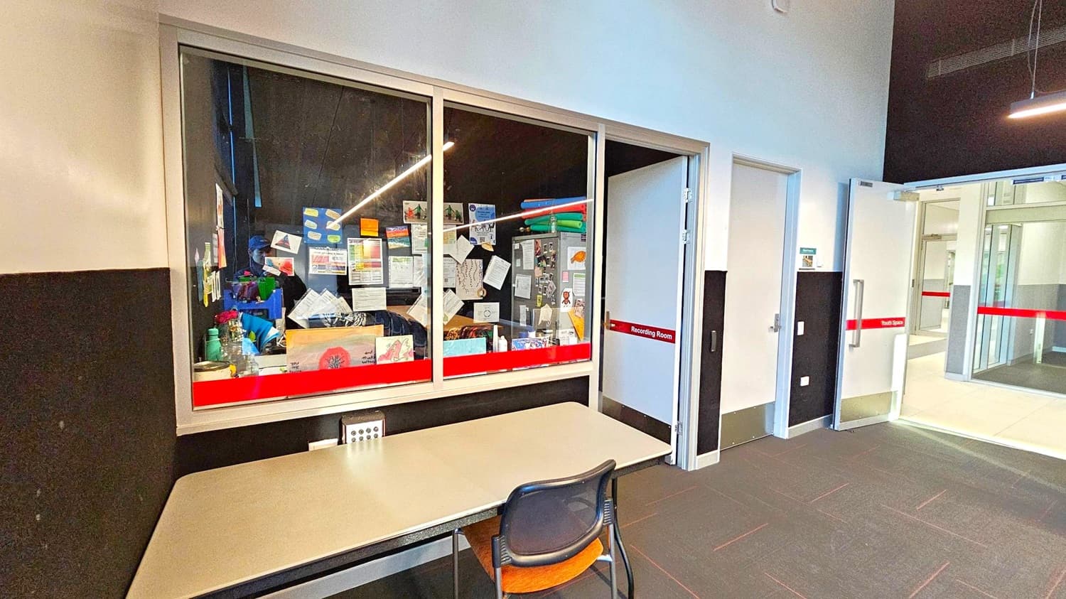 A room with a large window on the left, displaying various papers, drawings, and notices pinned inside. Below the window is a table with a chair. To the right, there are two doors; one is labelled "Recording Room" and the other leads to a corridor with glass doors. The floor is carpeted in a dark colour, and the ceiling is high with visible lighting fixtures.