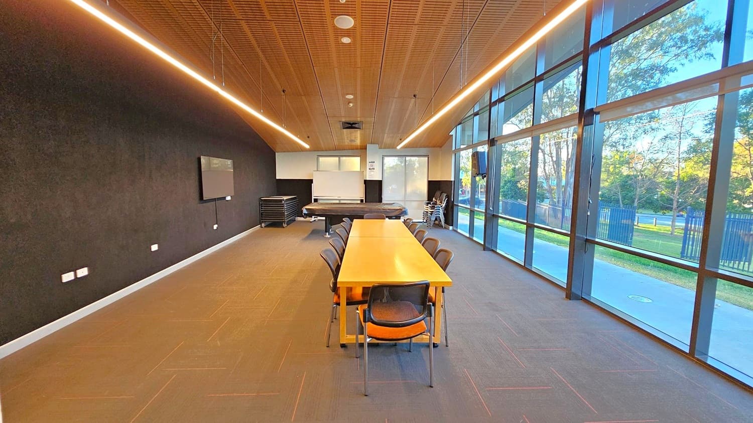 A modern conference room with a long wooden table in the centre, surrounded by chairs. The left wall is dark with a mounted screen, while the right side features large floor-to-ceiling windows offering a view of trees and a pathway outside. The back of the room has stacked chairs and a whiteboard. The ceiling is wooden with linear lighting, and the floor is carpeted in a neutral tone.