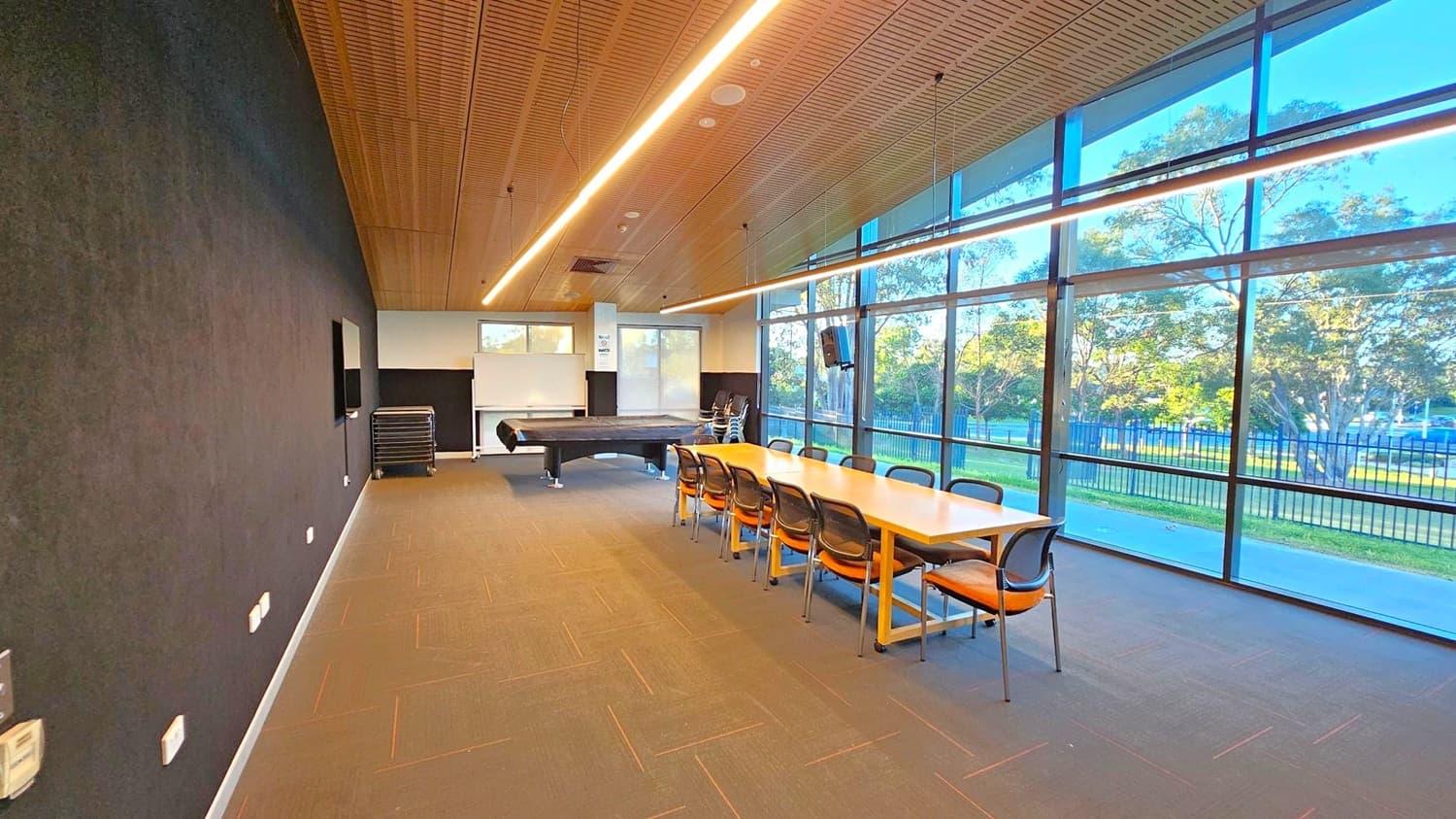 A room with a long wooden table and several chairs on the right side, next to large floor-to-ceiling windows showing trees and a fence outside. On the left, a dark wall with a mounted screen. At the far end, a pool table and a stack of chairs near a whiteboard. The ceiling is wooden with linear lights, and the floor is carpeted in a neutral tone.
