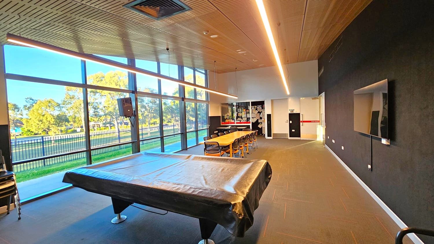 A room with large floor-to-ceiling windows on the left, offering a view of trees and a fence outside. In the centre, a long wooden table with chairs is positioned near a wall covered with notes and papers. A pool table with a cover is in the foreground. The right wall is dark with a mounted flat-screen TV. The ceiling is wooden with linear lights, and the floor is carpeted in a dark tone.