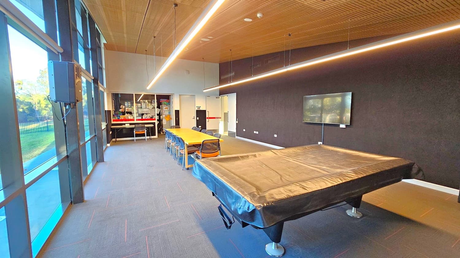 A room with a pool table covered in black fabric on the right. In the centre, a long wooden table with chairs is positioned. The left side features large windows with a view of greenery outside. The back wall has a display board and shelves with various items. A flat-screen TV is mounted on the right wall. The ceiling is wooden with linear lights, and the floor is carpeted in grey with subtle patterns.