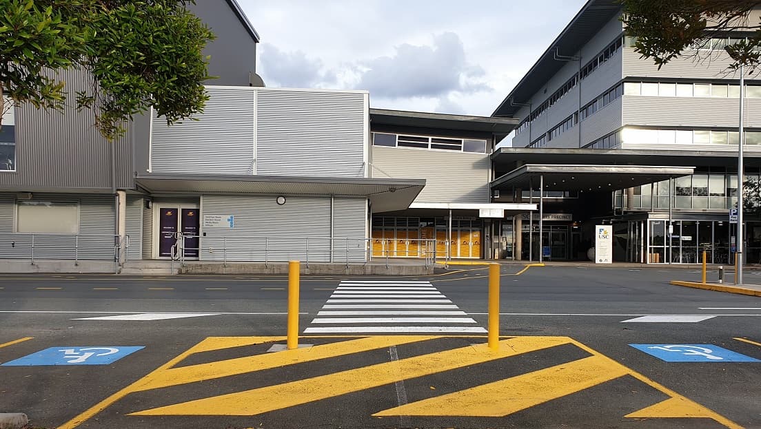 A large, industrial style building with a grey metal façade. On the left side are two purple-colored doors with descriptions on them, overlooking a walkway with pedestrian markings. In the center are yellow-patterned glass walls and a welcoming inscription on the door. The main entry is a well-lit grey façade with a roll-up banner and the USC inscription above a large glass door. Adjacent to it is a see-through glass seating area with signs in front.