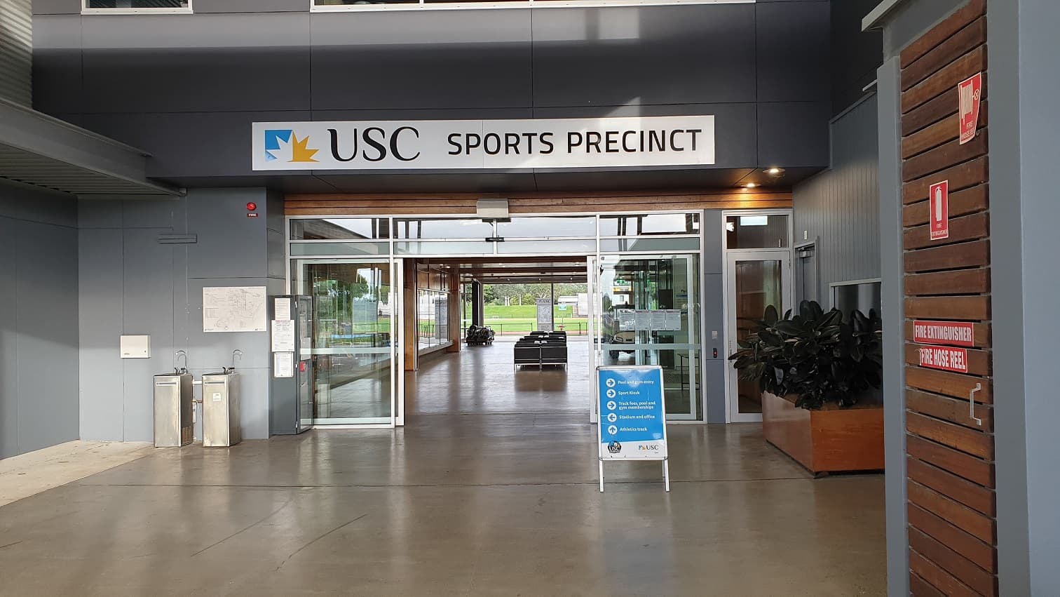 The main entrance has the USC sports precinct written on it and an automatic glass door that leads to a well-lit space with a row of chairs in the centre and a field at the end. Two sinks, a notice board, a fire alarm, and an infographic on the wall are on the left. A big rectangular-shaped pot plant holder, a sign with directions, and a small glass door facing the muster point by the wooden wall with directives on the right.