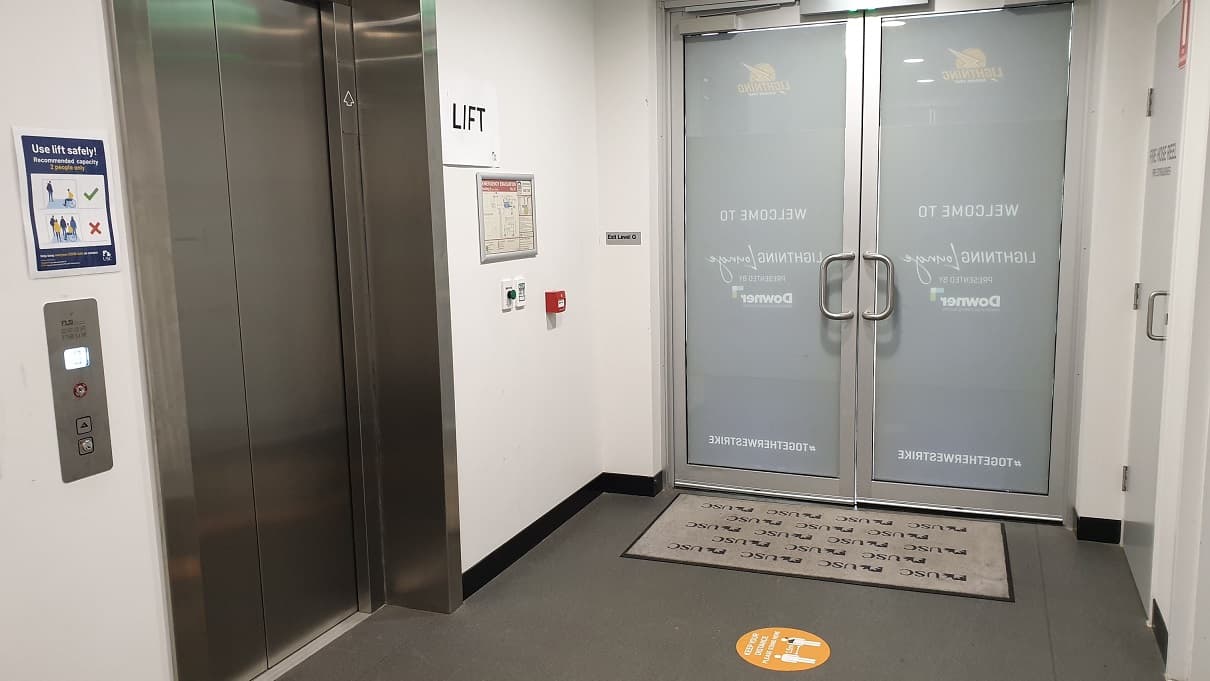 An elevator with instructions and signs around is positioned to the left of two glass, inward opening doors with a USC floor mat in front of the entrance.  To the right is white cupboard storing the fire hose reel.