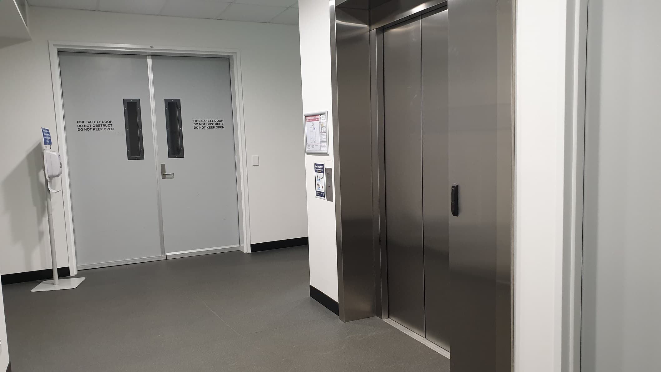 An elevator is positioned to the right and is stainless steel and well polished. Straight ahead are the emergency exit doors and a hand sanitiser dispenser sits to the left of the fire exit doors. The floor is grey vinyl.