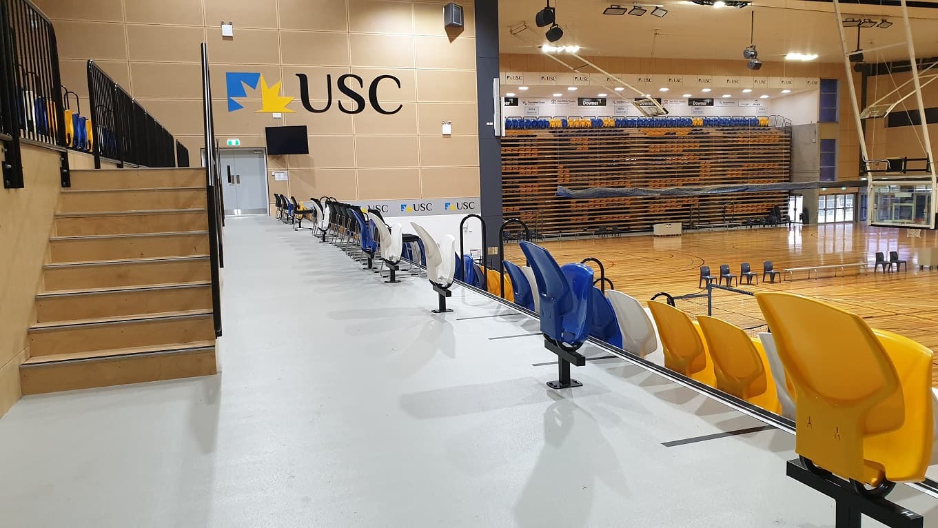 The stadium seating area is bordered by black metal railings and stairs lead up to the left to bleachers overlooking the arena with blue, yellow and white seats arranged in rows. Additional seating is to the left which leads down towards the stadium playing area further to the right.  A TV and a big USC logo are on the beige wall by grey emergency exit doors.