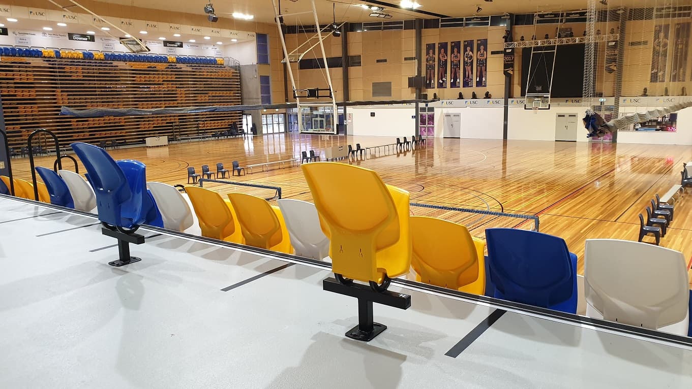 A view of the stadium playing area with blue, yellow and white seats in the foreground. A range of tables and chairs are set up on the stadium playing floor and additional seating is stacked in high rows and in storage to the left. On the back walls are a lineup of 10 players' photos on a beige wall. Rows of blue plastic seats and white benches, separated by white desks, overlook a white wall with covered decks, written words, and USC logos.