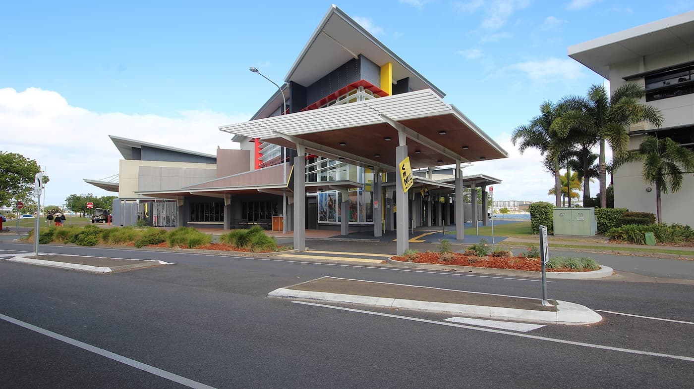 On the left, there is a curved road with a pavement and trees lining the side. On the right, there are modern buildings with large windows and some palm trees in front. In the middle, a building with a distinctive yellow vertical stripe and a slanted roof stands prominently. The background features more trees and parked cars. The foreground includes the road with white lane markings and a pedestrian crossing.