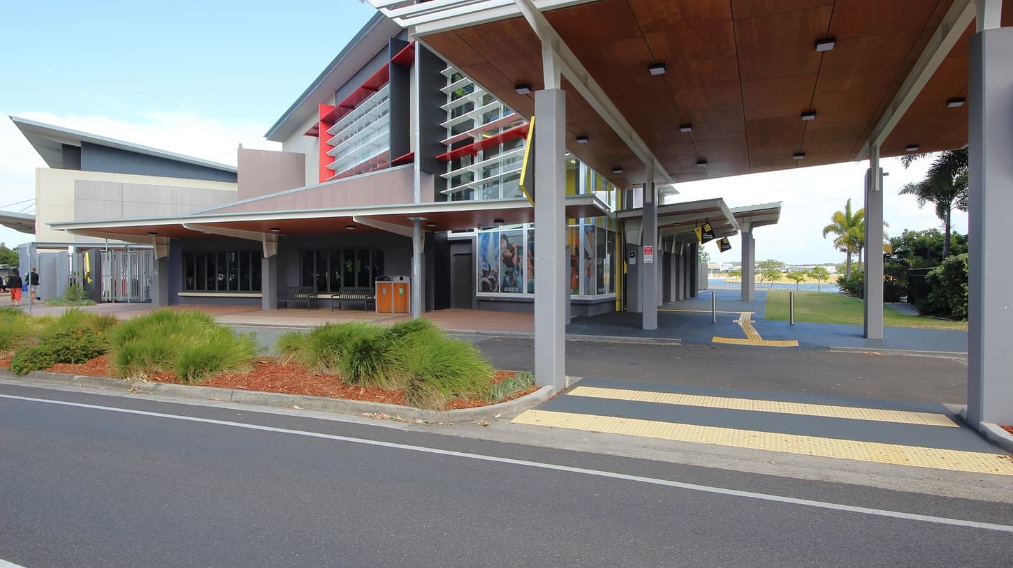 On the left, there is a modern building with large windows and a slanted roof, surrounded by landscaped areas with red mulch and shrubs. On the right, there is a covered walkway with a wooden ceiling and ceiling lights, leading to another building with palm trees nearby. In the middle, a prominent building with a yellow vertical stripe and large overhang extends towards the foreground. The foreground features a road with white lane markings and a pedestrian crossing with yellow tactile paving.
