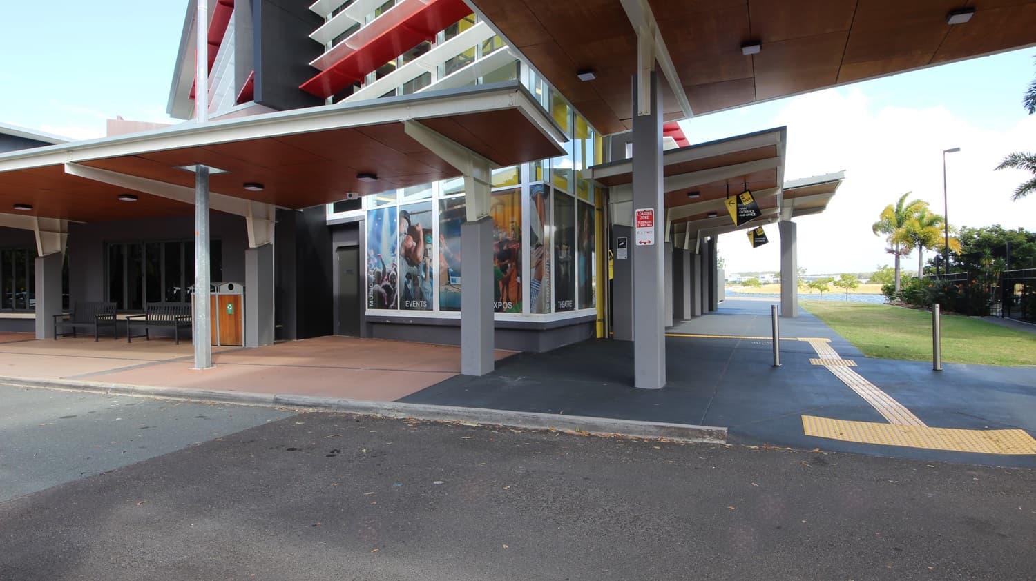 On the left, there is a building with large windows displaying posters and a wooden ceiling over a walkway. On the right, there is a covered area with a wooden ceiling and ceiling lights, leading to a pathway with yellow tactile paving. In the middle, a building with a yellow vertical stripe and red accents extends towards the foreground. The foreground features a paved area with a mix of dark and light grey flooring, and a small section of asphalt road.