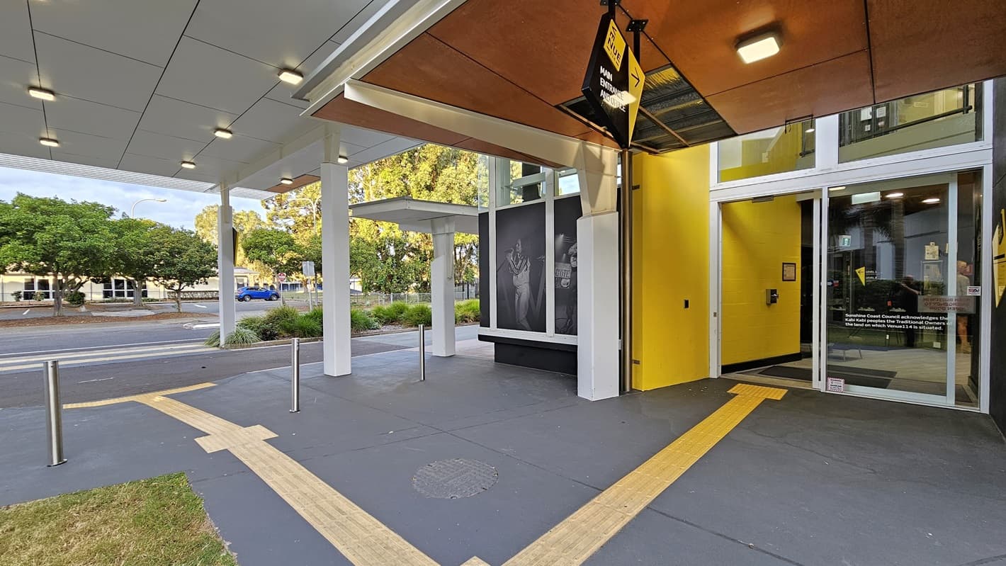 On the left, there is a covered walkway with a white ceiling and ceiling lights, leading to a building with large black-and-white posters. On the right, there is a glass entrance with a yellow wall and a door. In the middle, a yellow vertical stripe and a black overhang with a sign extend towards the foreground. The foreground features a dark grey floor with yellow tactile paving and a small patch of grass. The background includes trees and a road with parked cars.