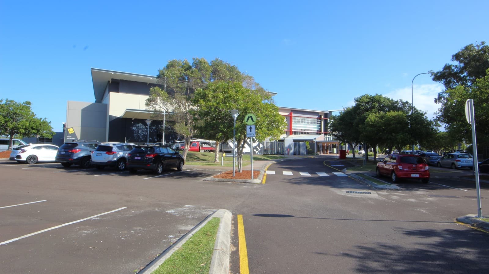 On the left, there are parked cars next to a building with a slanted roof and large windows. On the right, there are more parked cars and trees lining the area. In the middle, a building with red accents and multiple windows extends towards the background. The foreground features a paved road with white lane markings, a pedestrian crossing, and a small landscaped area with a tree and red mulch.
