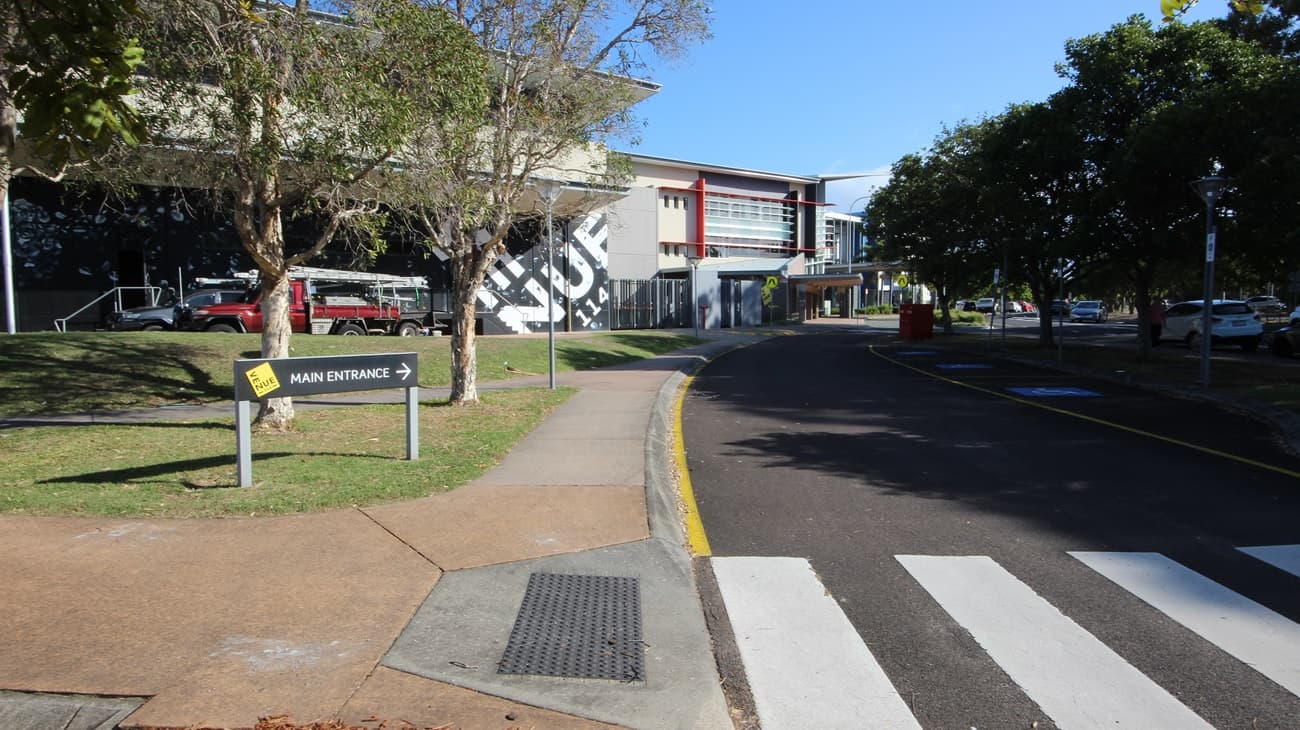On the left, there is a building with a large mural, a tree, and a sign pointing to the main entrance. On the right, there are trees and parked cars along a road. In the middle, a building with red and grey accents and multiple windows extends towards the background. The foreground features a pedestrian crossing with white stripes, a paved walkway, and a section of asphalt road with yellow lines.